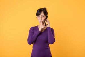 Mysterious Asian woman with purple shirt makes silence gesture presses index finger to lips shares secret with you holds mobile phone wears optical eyeglasses and warm jumper isolated over yellow wall photo
