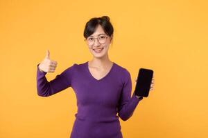 emocionado joven asiático mujer 30s vistiendo púrpura camisa y los anteojos demostración móvil teléfono pantalla, pulgares arriba, me gusta algo bien, recomendando teléfono inteligente solicitud, en pie terminado amarillo antecedentes foto