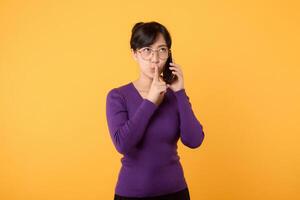 Mysterious Asian woman with purple shirt makes silence gesture presses index finger to lips shares secret with you holds mobile phone wears optical eyeglasses and warm jumper isolated over yellow wall photo