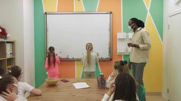Afrikaanse Amerikaans leraar in een masker geeft les een groep van gemaskeerd kinderen aan het leren engels. school- voor kinderen, onderwijs adolescenten, krijgen kennis, leren de taal. video