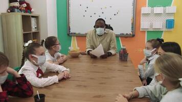 ein afrikanisch amerikanisch Lehrer und ein Gruppe von maskiert Kinder Studie im ein Klassenzimmer während ein Pandemie. Schule zum Kinder, Lehren Jugendliche, dazugewinnen Wissen, lernen das Sprache. video