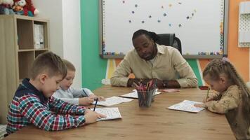 een Afrikaanse Amerikaans leraar geeft les een groep van kinderen naar trek terwijl zittend Bij een tafel in de klas. school- voor kinderen, onderwijs adolescenten, krijgen kennis, leren de taal. video