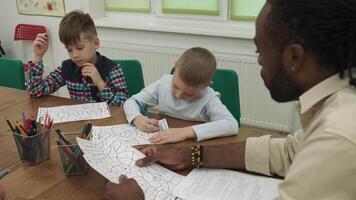 un africano americano profesor enseña un grupo de niños a dibujar mientras sentado a un mesa en el aula. colegio para niños, enseñando adolescentes, ganancia conocimiento, aprender el idioma. video