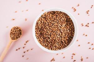 Close up of raw buckwheat in a bowl and wooden spoon on a pink background top view photo