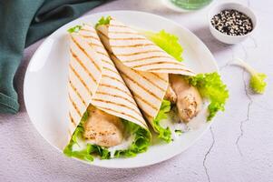 Grilled chicken fillet wrapped in tortilla with lettuce and sauce on a plate on the table photo