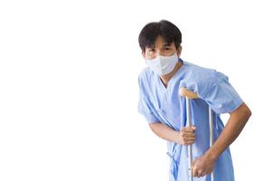 Asian male patient person in blue hospital gown holding onto crutch, indicative of injury or disability, capturing the essence of recovery or physical limitation while isolated white background. photo