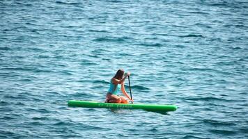 hav kvinna supera. silhuett av Lycklig positiv ung kvinna i blå bikini, surfing på grön supera styrelse genom lugna vatten yta. idyllisk solnedgång. aktiva livsstil på hav eller flod. långsam rörelse video