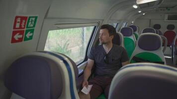 Young man traveling on an empty Italian train with colorful seats using his smartphone and holding tickets in hand on a sunny summer day in Rome, Italy. Tourist rides on railway checks route on phone. video