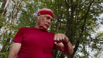 Senior corredor homem dentro parque usando inteligente assistir, rastreamento distância, verificação pulso depois de ginástica exercite-se video