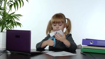 Child schoolgirl learns lessons at home sitting at table cutting with scissors shapes out of paper video