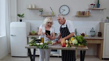 Vegan senior couple cooking salad with raw vegetables. Looking on digital tablet for online recipe video