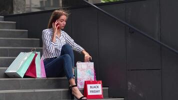 meisje zittend Aan trap met Tassen pratend Aan smartphone over uitverkoop in boodschappen doen winkelcentrum in zwart vrijdag video