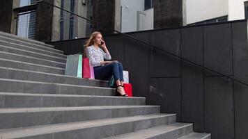 Girl sitting on stairs with bags talking on mobile phone about sale in shopping mall in Black Friday video