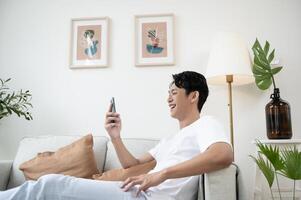 contento asiático hombre sentado en sofá utilizando teléfono inteligente en vivo habitación a hogar, relajarse hora y estilo de vida concepto foto