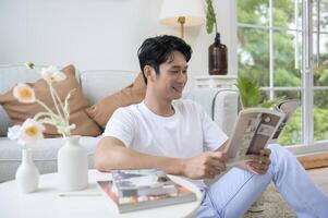 contento asiático hombre sentado en sofá leyendo un libro en vivo habitación a hogar, relajarse hora y estilo de vida concepto foto