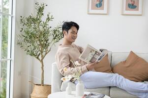 Happy asian man sitting on couch  reading a book in living room at home, relax time and lifestyle concept photo