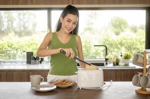 asiático mujer preparando café y brindis un pan para desayuno a el cocina mesa en el Mañana foto