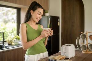 asiático mujer preparando café y brindis un pan para desayuno a el cocina mesa en el Mañana foto