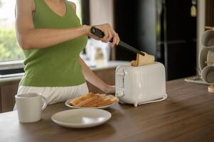 asiático mujer preparando café y brindis un pan para desayuno a el cocina mesa en el Mañana foto