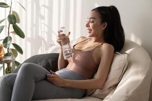 Young asian woman in sportswear drinking pure water from glass in living room at home photo