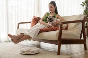 Young asian woman relaxing with pug dog in living room while Robotic vacuum cleaner working photo