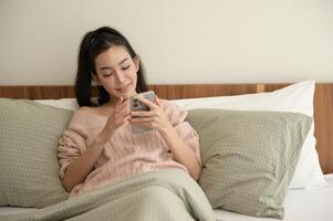 joven asiático mujer utilizando inteligente teléfono acostado en cama foto