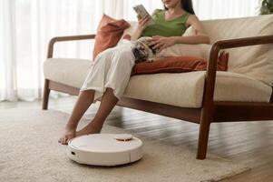 Young asian woman relaxing with pug dog in living room while Robotic vacuum cleaner working photo