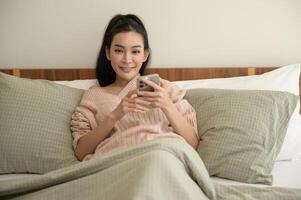 joven asiático mujer utilizando inteligente teléfono acostado en cama foto