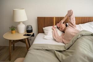 dormido asiático mujer torneado apagado alarma en teléfono inteligente mientras siendo despertar arriba en el Mañana foto