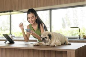 joven asiático mujer trabajando con tableta y disfrutando con su perro en el cocina a hogar foto