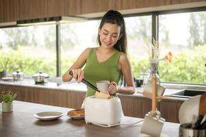 asiático mujer preparando café y brindis un pan para desayuno a el cocina mesa en el Mañana foto