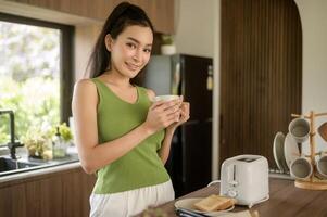 asiático mujer preparando café y brindis un pan para desayuno a el cocina mesa en el Mañana foto