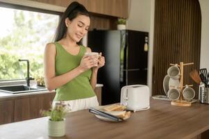 asiático mujer preparando café y brindis un pan para desayuno a el cocina mesa en el Mañana foto