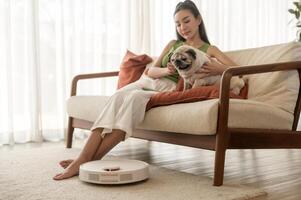 joven asiático mujer relajante con doguillo perro en vivo habitación mientras robótico vacío limpiador trabajando foto