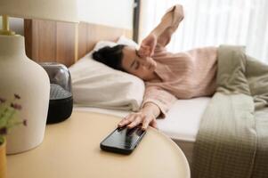 dormido asiático mujer torneado apagado alarma en teléfono inteligente mientras siendo despertar arriba en el Mañana foto