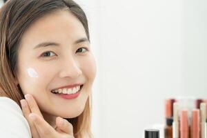 Beautiful Asian woman sit in front of a mirror. face of a healthy woman applying cream and makeup. Advertisement for skin cream, anti-wrinkle, baby face, whitening, moisturizer, tighten pores serum photo