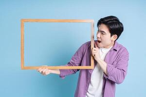 Young Asian man holding photo frame on blue background