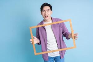 Young Asian man holding photo frame on blue background