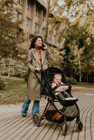 joven mujer con linda bebé niña en bebé paseante utilizando móvil teléfono a el otoño parque foto