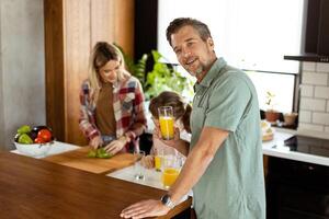 Joyful man with a glass of orange juice while a woman and child prepare food in a cozy kitchen photo