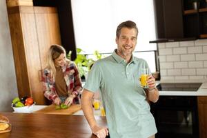 Smiling man holds a glass of orange juice while two women cook in a bright kitchen photo