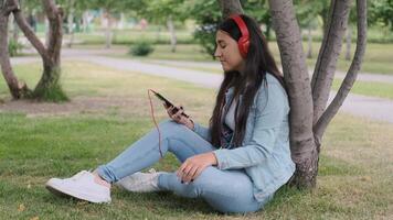 Happy girl with dark hair and jeans clothes is listening to music with headphones while sitting near a tree in the park. video