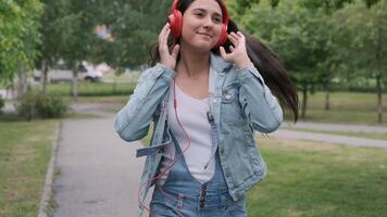 lindo alegre menina com Sombrio cabelo e uma jeans Jaqueta dançando caminhando dentro a parque com fones de ouvido video