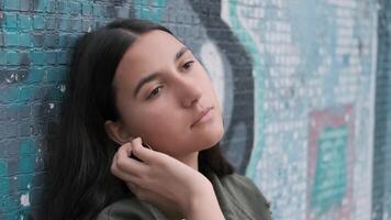 young brunette girl with long hair holds her hand through her hair and is sad leaning on the wall with graffiti video