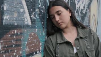 portrait de une Jeune triste brunette branché fille avec marron yeux et longue cheveux près une mur avec graffiti video