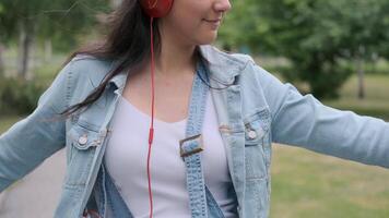 gracioso alegre contento niña en mezclilla ropa bailando en el parque durante el día escuchando a música con auriculares. divertido humor. de cerca video