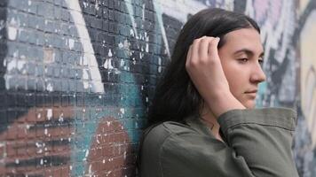 Young beautiful pensive brunette girl with long hair stands near the wall with graffiti and runs her hand through her hair. video