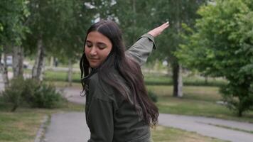 alegre hipster menina dentro verde roupas é caminhando e dançando dentro a parque para a música enquanto segurando uma portátil alto falante dentro dela mão. Câmera rotação video