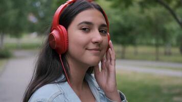 rolig glad Lycklig flicka i jeans kläder dans i de parkera nära de träd i de eftermiddag lyssnande till musik i hörlurar. roligt humör. porträtt video