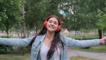 alegre menina com Sombrio cabelo dentro fones de ouvido anda em em a rua e escuta para música dentro uma Boa humor video